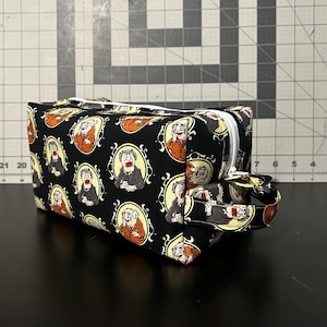 May include: A rectangular fabric toiletry bag with a black background featuring a repeating pattern of two elderly men in oval frames. The bag has a white zipper closure along the top and a small fabric handle on the side for easy carrying and storage.