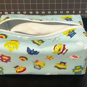 Peut inclure: Une trousse de toilette rectangulaire bleu clair avec une fermeture &eacute;clair blanche et des poign&eacute;es. Le sac est recouvert d'un motif d'aliens verts, d'&eacute;toiles et d'autres personnages. Le sac est ouvert, r&eacute;v&eacute;lant un int&eacute;rieur blanc.