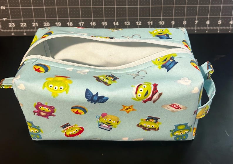 Peut inclure: Une trousse de toilette rectangulaire bleu clair avec une fermeture &eacute;clair blanche et des poign&eacute;es. Le sac est recouvert d'un motif d'aliens verts, d'&eacute;toiles et d'autres personnages. Le sac est ouvert, r&eacute;v&eacute;lant un int&eacute;rieur blanc.