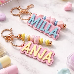 May include: Two personalised keyrings, one with "MILLA" in light blue and the other with "ANJA" in yellow, on pink nameplates. Both have gold-coloured rings and are decorated with colourful beads. The background features pastel-coloured accessories.