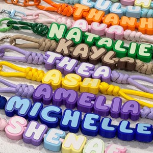May include: A collection of colourful lanyards featuring custom names spelled out in chunky, block-style plastic letters. Each lanyard is attached to a metal lobster clasp and includes a matching cord in various shades like orange, green, purple, and blue.