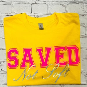 May include: Yellow t-shirt with the words "SAVED Not Soft" in pink and silver lettering.