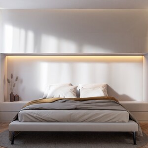 Puede incluir: Una cama con un marco blanco, un edred&oacute;n gris y dorado y dos almohadas blancas. La cama est&aacute; en un dormitorio moderno con una pared blanca y una l&aacute;mpara sobre la cabecera.