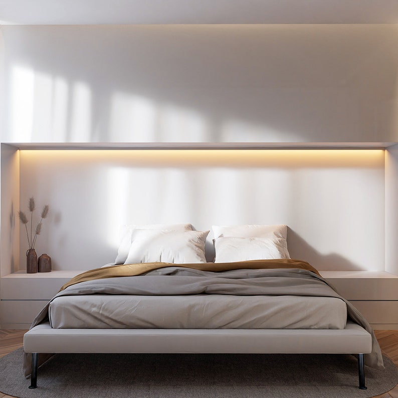 Puede incluir: Una cama con un marco blanco, un edred&oacute;n gris y dorado y dos almohadas blancas. La cama est&aacute; en un dormitorio moderno con una pared blanca y una l&aacute;mpara sobre la cabecera.