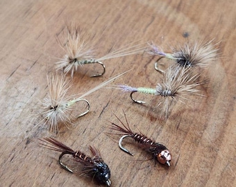 May include: Five different fly fishing lures with brown, black, and white feathers. The lures have silver hooks and are arranged on a wooden surface.