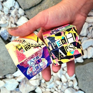 May include: Two small, colorful wooden cubes with abstract designs and text. The cubes feature a mix of bright colors, including yellow, pink, blue, and green. The text on the cubes includes "SUPER" and "ABS OF STEEL".