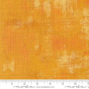 May include: A square of fabric with a textured, abstract design in shades of yellow, orange, and cream. The fabric has a distressed, painted appearance with visible brushstrokes and a grid-like pattern. The bottom edge features a ruler with inch measurements.