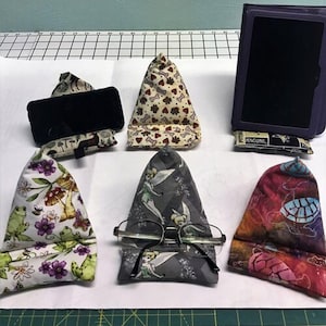 Puede incluir: Una colección de soportes de tela para tabletas y teléfonos con varios diseños. Los diseños incluyen huesos de perro, ranas, setas, Campanilla y tortugas. Un soporte sostiene unas gafas. Los soportes están diseñados para sostener dispositivos para su uso con manos libres.