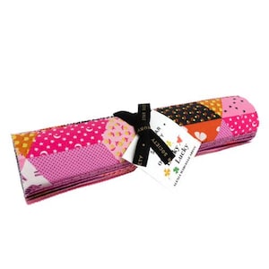 Puede incluir: Una pila de cuadrados de tela rosa, naranja y negro sobre un fondo blanco. La tela está estampada con estrellas, lunas y otras formas geométricas. La tela está atada con una cinta negra y una etiqueta que dice "Star Society - Happy Lucky".