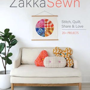 May include: A cream-colored sofa with three decorative pillows. The pillows are orange, yellow, and beige with various patterns. A framed circular quilt hangs on the wall above the sofa. The book "ZakkaSewn" is visible.