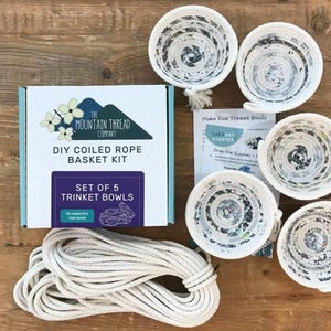 May include: A DIY coiled rope basket kit by The Mountain Thread Company, including a box labeled Set of 5 Trinket Bowls, a coil of thick white rope, five finished black and white patterned coiled rope bowls, and an instructional paper sheet.