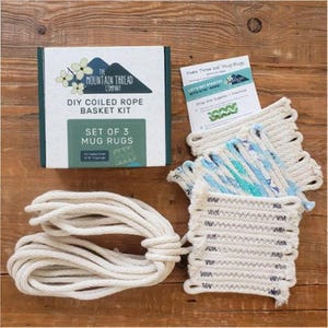 May include: A DIY coiled rope basket kit for three mug rugs, including a white box with text, a printed instruction card, a coil of thick off-white cotton rope, and three finished square mug rugs featuring blue, green, and white zigzag patterns.