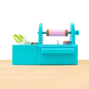 Puede incluir: Un organizador de costura azul turquesa con un cajón y un soporte para carretes. El soporte para carretes tiene un carrete de hilo rosa con una etiqueta marrón que dice "Aurifil Made in Italy 100% Cotton".