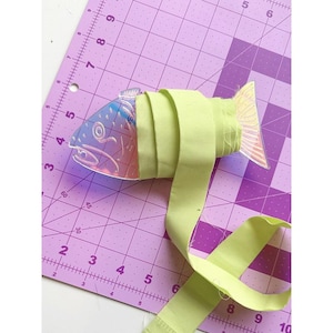 Może przedstawiać: Przezroczysty, opalizujący przedmiot w kształcie ryby jest owinięty jasnozielonym paskiem materiału. Ryba ma wytrawione detale i przezroczystą płetwę ogonową. Materiał jest owinięty wokół ciała ryby i rozciąga się na zewnątrz.