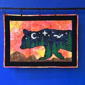 May include: A quilted wall hanging featuring a bear silhouette filled with a night sky design. The bear is dark blue, with a crescent moon, stars, and a flying bird. The background is a sunset of orange, pink, and yellow.