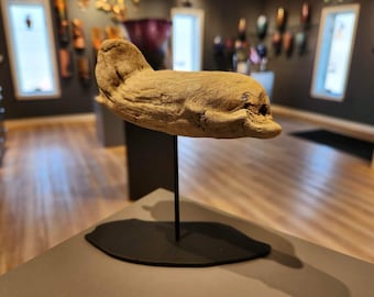 May include: A driftwood sculpture of a dolphin, painted white, mounted on a black stand. The sculpture is displayed on a light wood surface.