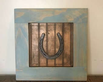 Puede incluir: Una decoración de pared de madera rústica con un marco pintado de azul y un panel de madera marrón con una herradura de metal.