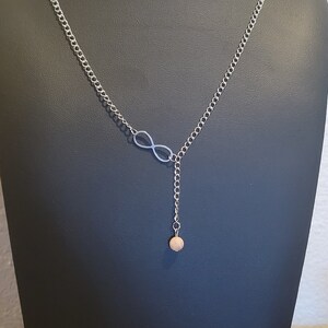 May include: Silver-toned necklace featuring an infinity symbol pendant. The chain leads to a dangling strand ending with a small, round, light brown bead. The necklace is displayed on a black mannequin.