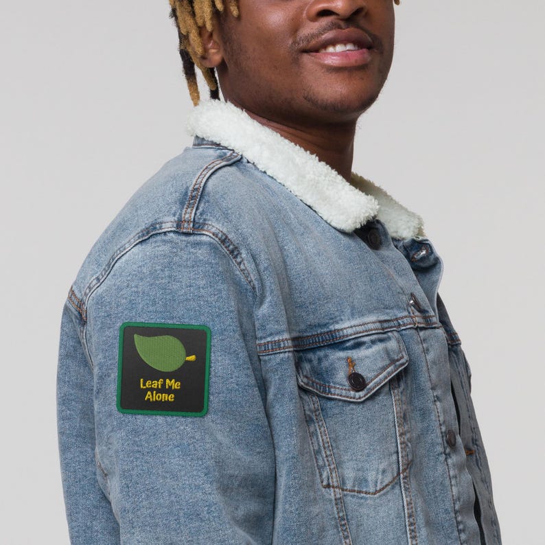 May include: A light blue denim jacket with a white sherpa collar. A green and black patch on the jacket reads "Leaf Me Alone" with a leaf graphic. The jacket has a button closure and a chest pocket.