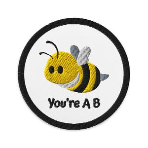 May include: Embroidered patch featuring a yellow and black cartoon bee with gray wings. The bee is inside a white circle with a black border. The text "You're A B" is below the bee.