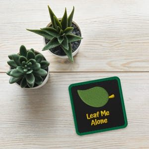 May include: A square patch with a green border and the words "Leaf Me Alone" in yellow. The patch features a green leaf design on a black background. Two small potted succulents are also in the image.
