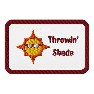 May include: Embroidered patch with a white background and a maroon border. The patch features a yellow sun wearing sunglasses, with the words "Throwin' Shade" in maroon lettering. The patch is rectangular with rounded corners.