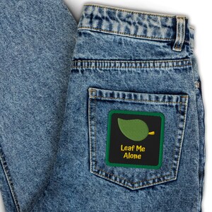 May include: Light blue denim jeans with a back pocket featuring a square patch. The patch has a green border and a black background with a green leaf design and the text "Leaf Me Alone" in yellow.