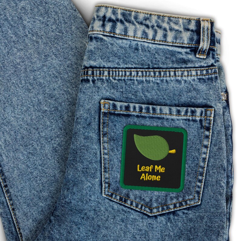 May include: Light blue denim jeans with a back pocket featuring a square patch. The patch has a green border and a black background with a green leaf design and the text "Leaf Me Alone" in yellow.