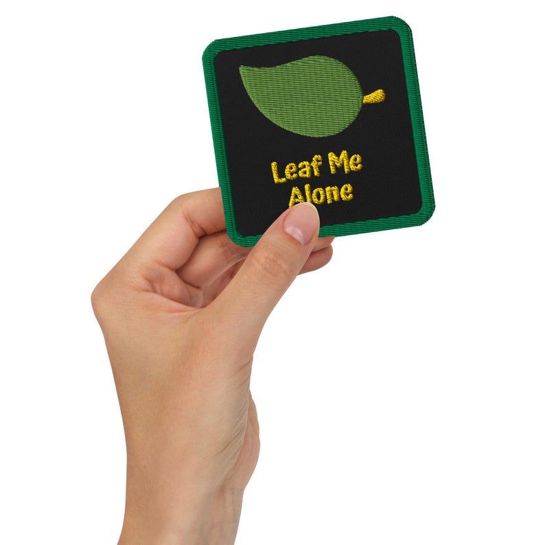 May include: Embroidered patch with a green leaf design and the text "Leaf Me Alone" in yellow. The patch has a black background with a green border. The patch is square-shaped.