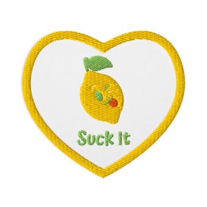 May include: Embroidered heart-shaped patch with a yellow border and white center. A yellow lemon with a green leaf and red tongue is in the center. The text "Suck It" is embroidered in green below the lemon.