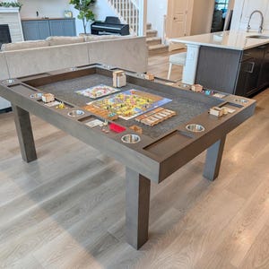 May include: A large, rectangular wooden game table with a gray finish. The table has built-in cup holders and recessed areas for game pieces. A board game is set up on the felt-lined playing surface, with various game pieces and accessories.