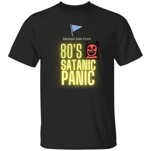 Puede incluir: Camiseta negra con un gráfico amarillo que dice "Marked Safe From 80's Satanic Panic". Un diablo de dibujos animados rojo con cuernos está sobre el texto. Una bandera azul está en la esquina superior izquierda.