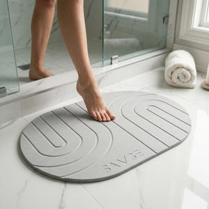 May include: An oval-shaped light gray bath mat featuring a textured surface with embossed U-shaped lines and the word SAVGE debossed on the bottom right. The mat is positioned on a white tiled floor in front of a glass shower enclosure.