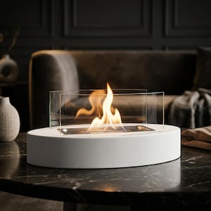 May include: A tabletop fireplace featuring a smooth, oval-shaped white concrete base with a central rectangular metal burner. Two curved, transparent glass panels are positioned vertically on the base, surrounding a bright, flickering orange flame.