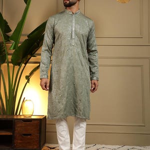 May include: A sage green kurta with detailed embellishments, worn with white trousers. The long-sleeved tunic has a mandarin collar and decorative elements. The outfit is completed with brown sandals, creating a stylish ensemble.