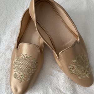 May include: A pair of gold-colored slip-on shoes. Each shoe features an embroidered crest design in a lighter gold thread. The shoes have a rounded toe and a low heel, suitable for casual wear.