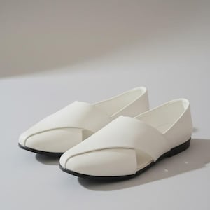 May include: A pair of white slip-on flats featuring a textured upper with a distinctive overlapping crossover design at the vamp, a closed rounded toe, and a thin black outsole. The shoes have a low-cut silhouette with a clean, solid white finish.