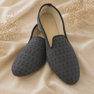 May include: A pair of dark gray slip-on loafers featuring a repeating pattern of small black diamonds with centered gold-toned dots. The shoes have a rounded toe, a low profile, and a dark trim along the collar, with a light-colored interior lining.