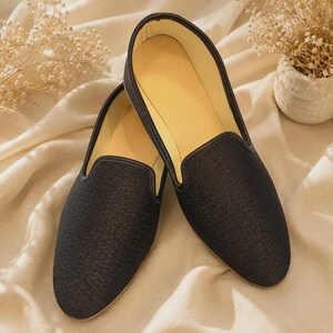May include: A pair of black slip-on loafers featuring a textured fabric upper with a subtle geometric pattern. The shoes have a rounded toe, a low-cut collar with black trim, and a light beige interior lining, presenting a clean and simple design.