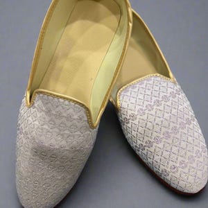 May include: A pair of cream-colored slip-on shoes with a geometric pattern. The shoes have gold trim along the edges and a gold interior. The soles are red. The shoes are likely for formal occasions.
