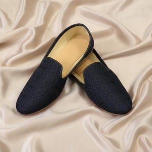 May include: A pair of navy blue slip-on shoes with a geometric pattern. The shoes have a light brown interior and are set against a backdrop of flowing, cream-colored fabric. The shoes appear to be made of a soft, textured material.