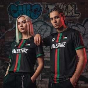 Puede incluir: Camisetas negras con la palabra "PALESTINA" en blanco, con rayas rojas y verdes y la bandera palestina. Las camisetas tienen ribetes verdes y las llevan dos personas delante de un muro de ladrillo cubierto de grafitis.