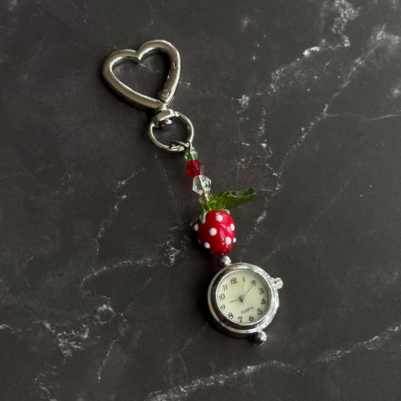 May include: A silver-toned keychain with a heart-shaped clasp, adorned with a red and white strawberry charm, green leaf, and a small quartz watch. The watch face displays the time. The charm is attached with colorful beads.