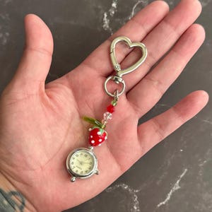 May include: A silver-toned quartz watch with a cream-colored face, attached to a heart-shaped clasp. The watch is adorned with a red strawberry charm with white dots, green leaves, and red and clear beads. The watch is approximately 1 inch in diameter.