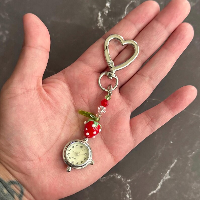 May include: A silver-toned quartz watch with a cream-colored face, attached to a heart-shaped clasp. The watch is adorned with a red strawberry charm with white dots, green leaves, and red and clear beads. The watch is approximately 1 inch in diameter.