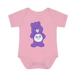 May include: A pink baby onesie with a purple Care Bear wearing a blue and pink lollipop on its belly.