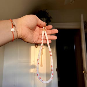 May include: A wristlet keychain made of white seed beads arranged in a tubular pattern, featuring small orange flower motifs spaced evenly throughout the length. The beaded loop is attached to a gold-toned metal ring with a tan leather connector.
