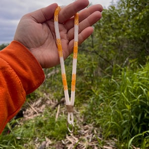 May include: A beaded lanyard in white and orange sections. The lanyard has a small metal ring and a leather-like piece. The background is a natural outdoor setting with green foliage.