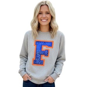May include: A gray sweatshirt featuring a large, blue sequined letter "F" outlined in orange. The sweatshirt is a crew neck style and is made of a soft material. The design is centered on the front of the shirt.
