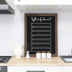 May include: A black chalkboard with a wooden frame hangs on a white wall. The chalkboard has the text "What's for dinner?" written on it and is divided into seven sections, one for each day of the week. The sections are labeled with the first letter of each day of the week: S, M, T, W, T, F, S.